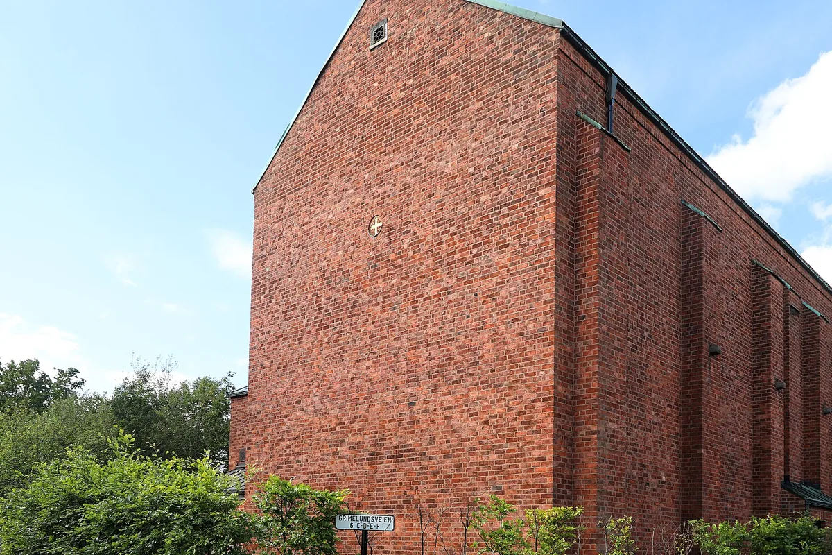The mausoleum from the street corner: a tall windowless building of red brick with a green copper roof, trees and hedges along its foot.