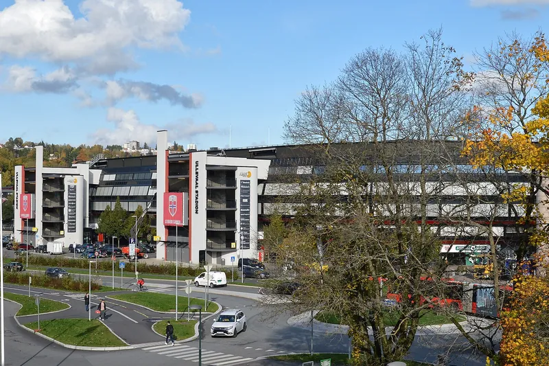 Ullevaal Stadion seen from the street on an autumn day: the long stand with its car park and entrances, trees in front.