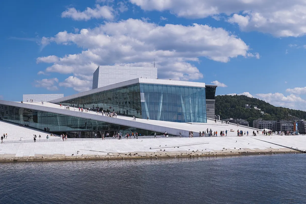The white Oslo Opera House on the waterfront, with people walking up its sloping roof under a blue sky.